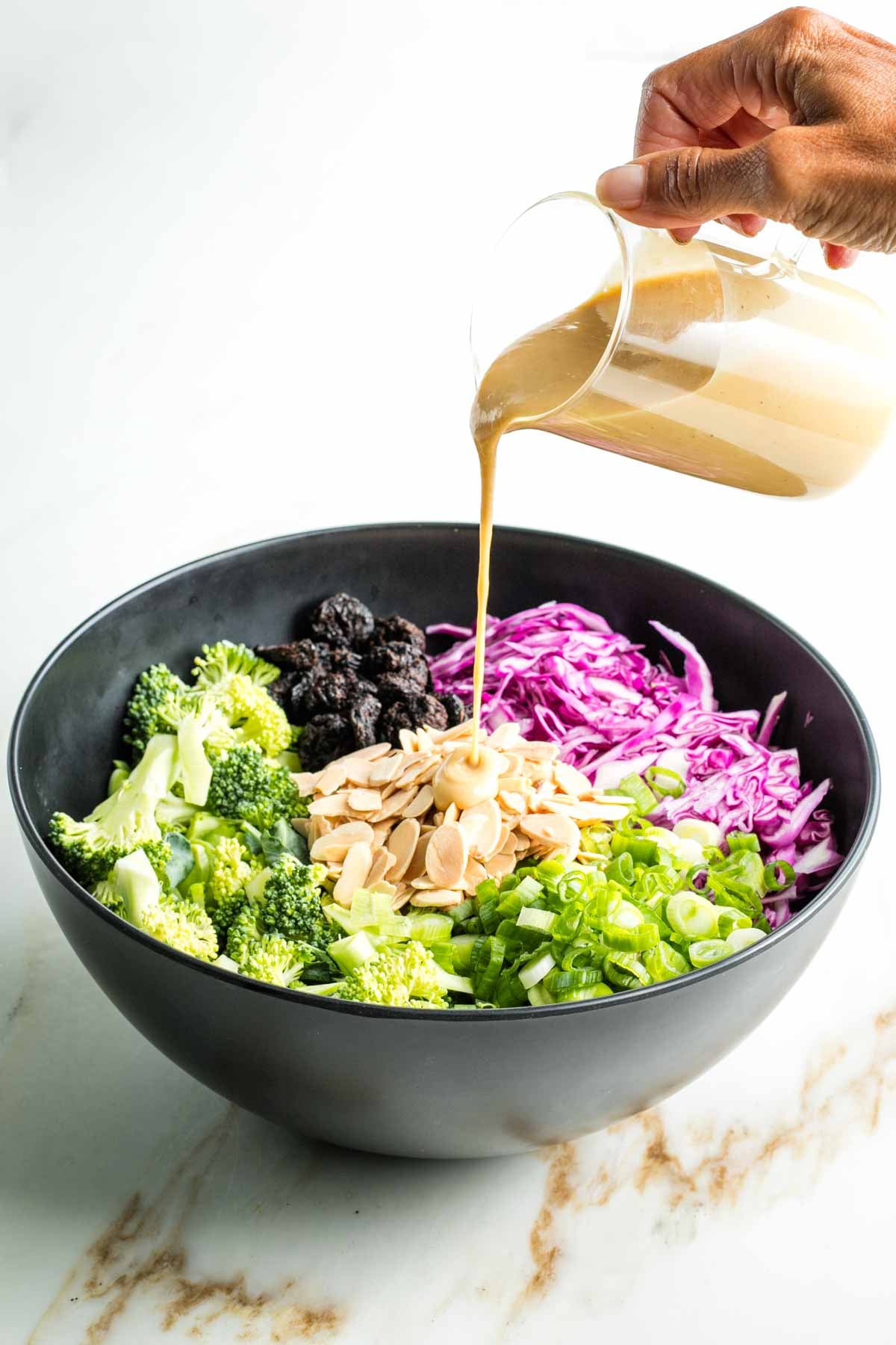 Pouring miso dressing over raw broccoli, cabbage, scallions plus almonds and dried cherries in a bowl. 