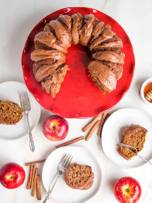 Apple Bundt Cake, drizzled with chocolate sauce on a red pedestal. Slices of cake on white plates, plus whole apples and cinnamon sticks surround the pedestal.