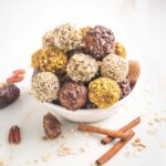 White bowl with pumpkin energy balls stacked up. They are coated with different nuts and seeds. In the foreground are dates, oats, cinnamon sticks and whole pecans.