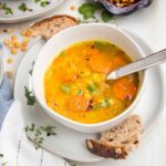 White bowl filled with yellow split pea soup with carrots and fresh herbs. Crusty bread pieces on the plate holding the soup bowl.