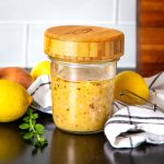 Glass jar with wooden lid filled with salad dressing, surrounded by fresh lemons, a shallot and some fresh herbs.