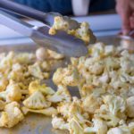 Cauliflower Florets sprinkled with spices on a baking sheet. Metal tongs are tossing the florets on the baking tray.