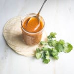 Jar of red enchilada sauce with a spoon in it on top of a stack of tortillas with some cilantro on the side.