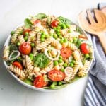 Large white bowl filled with cherry tomatoes, fusilli pasta, green peas, chick peas, sunflower seeds with a striped blue napkin on the side.