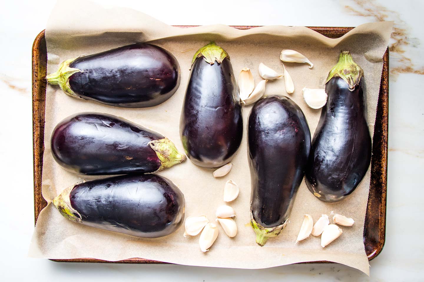 Halved eggplant and garlic cloves with their skins on a parchment lined baking tray.