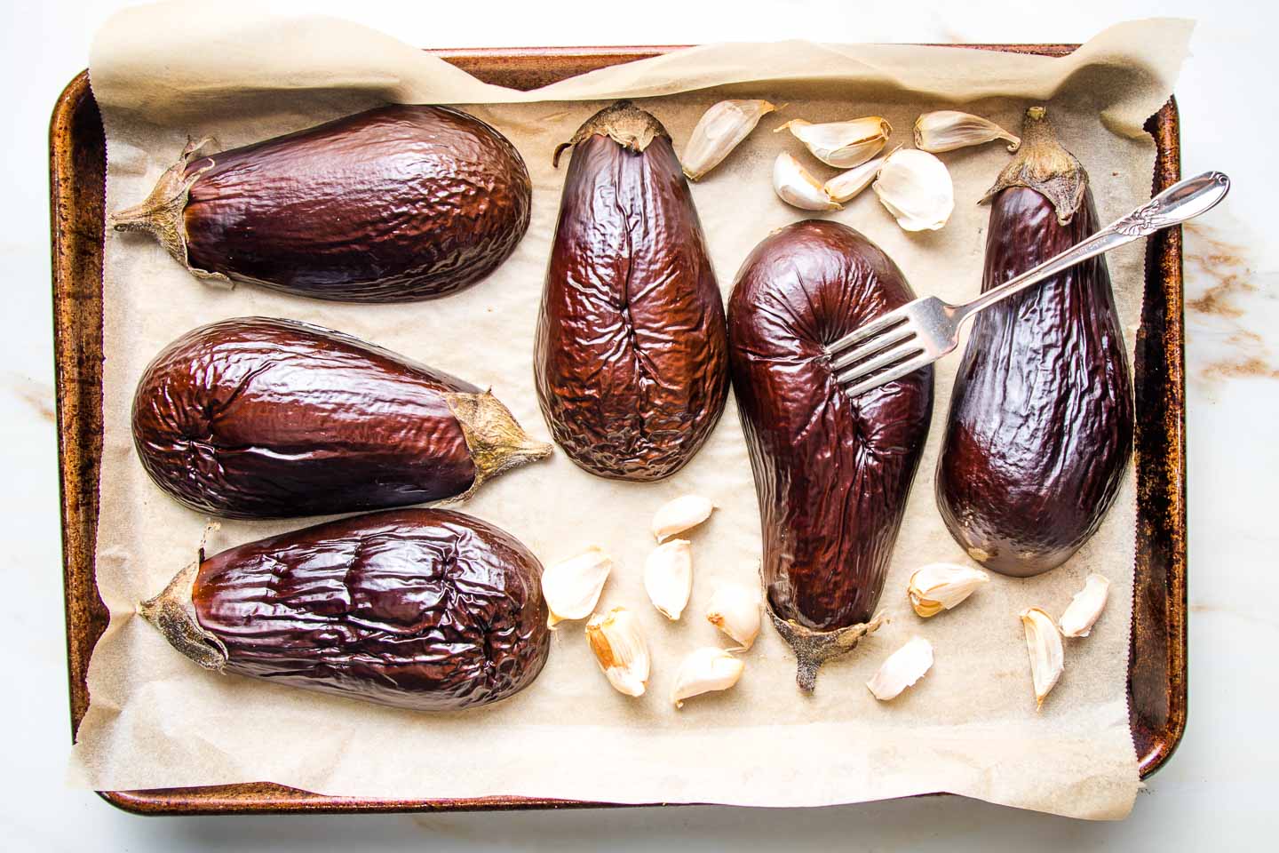 Halved eggplant roasting on a parchment lined baking tray with garlic cloves in their skins.