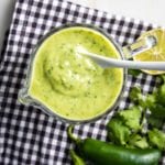 Black and white checkered cloth with a glass pitcher filled with avocado cilantro lime dressing, plus cilantro, jalapeno and a lime on the side.