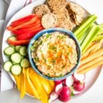 Blue bowl filled with Roasted Eggplant Dip sprinkled with parsley and paprika on top of a white plate with brightly colored veggies for dipping.