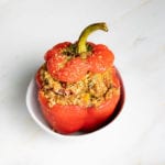Red bell pepper that is stuffed with quinoa and lentils plus italian spices, with it's top back on after cooking sitting inside a white bowl.
