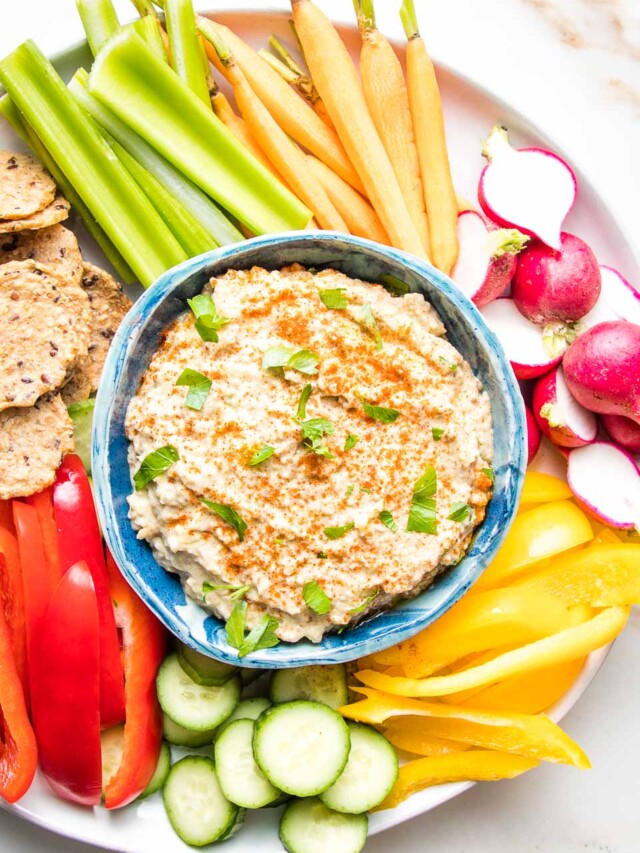 Baba Ganoush in a blue bowl sprinkled with paprika and fresh parsley with crudites of brightly colored radish, bell peppers, celery, carrots and crackers.