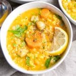 White bowl filled with thick soup. Bright yellow broth with chickpeas, rice, carrots, lemon wedge and sprinkled with chopped parsley, salt and pepper.