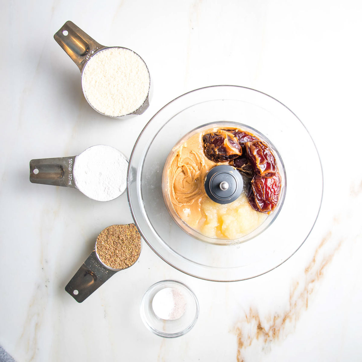 Dates, applesauce and peanut butter in the bowl of a food processor plus measuring cups with almond flour, cassava flour, ground flax, salt and baking soda. 