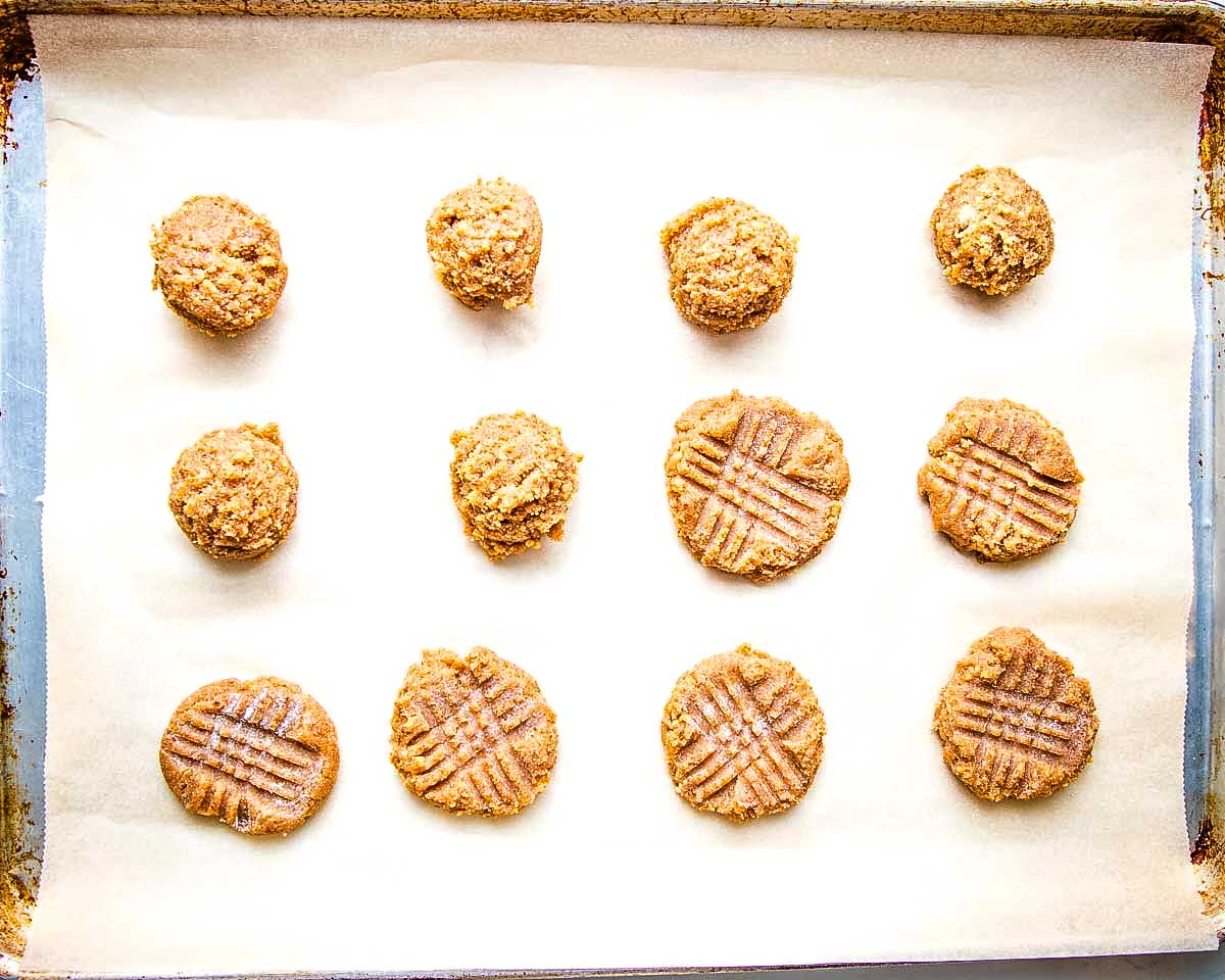 Peanut butter cookies dropped onto a baking sheet, some with crosshatch pattern fork marks. 