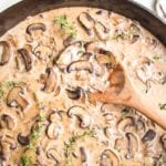 Close up of cast iron skillet filled with creamy vegan sauce, loaded with sliced mushroom, fresh thyme and a wooden spoon stirring it.