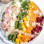 Shredded rainbow salad on a round rimmed white plate: a layer of arugula covered with shredded radish, green cabbage, beet leaves, roasted delicata squash, shredded carrots, purple cabbage and red beets.