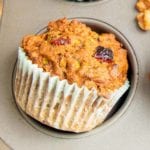 close up of a morning glory muffin, showing a dried cranberry and some shredded carrots. The muffin has a light blue paper liner sitting just outside the pan it was baked in.