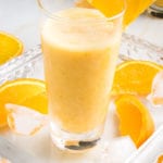 Glass filled with a frothy Orange Smoothie, with a wedge of orange on the side of the glass, more orange wedges on the glass serving tray along with some ice cubes.