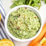 Close up of carrot top pesto in a small white bowl, sprinkled with hemp seeds and crushed red pepper. You can see glimpses of carrots, arugula and lemons surrounding the bowl.