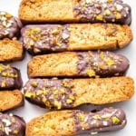 Biscotti, half dipped in dark chocolate and sprinkled with nuts, sea salt and orange zest on a white platter.