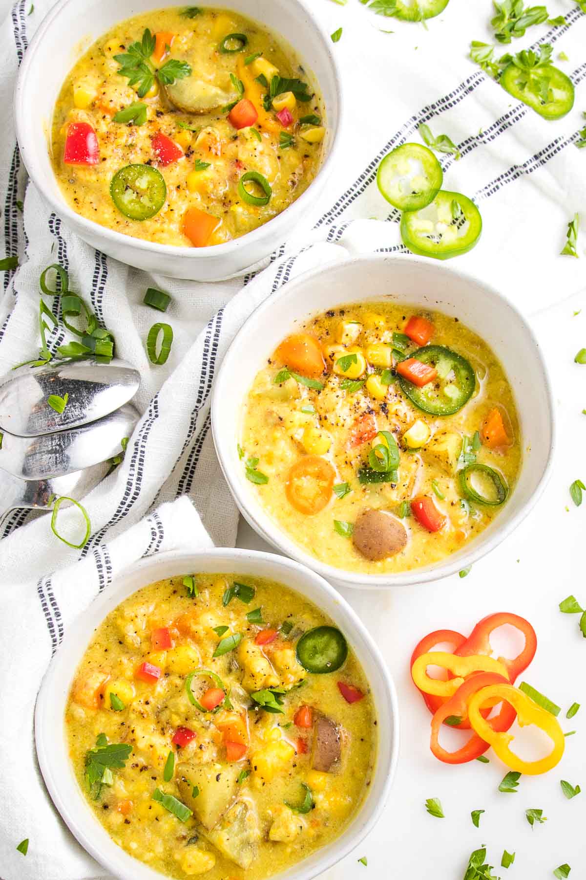 Three white bowls, filled with corn chowder, and chunky colorful veggies. There is a white tea towel with dark thin stripes and some scallion slices and red and yellow pepper slices on the counter.