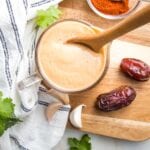 Small glass pitcher filled with orange creamy salad dressing, there are a couple of dates, a few cloves of garlic, some sprigs of cilantro and a small dish of chipotle powder surrounding the dressing that's on top of a wooden cutting board.