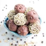 Small blue bowl filled with chocolate cookie dough balls. Some have a coating of hemp seeds on the outside. There are mini chocolate chips and bits of rolled oats scattered around the bowl on a white marble countertop.
