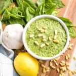 Close up of a bowl of pesto sprinkled with pine nuts...there is a lemon, a head of garlic, some fresh basil leaves and some pine nuts flanking the pesto bowl on a wooden board.