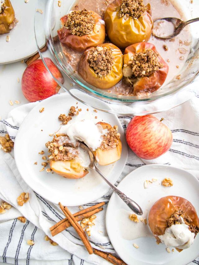 White plate with a baked apple, stuffed with oatmeal mixture cut in half and topped with vanilla ice cream. There is another plate on the bottom right with a whole baked apple, a glass pie plate iwth additional baked apples and the background is scattered with cinnamon sticks, red uncooked apples and crumbs.