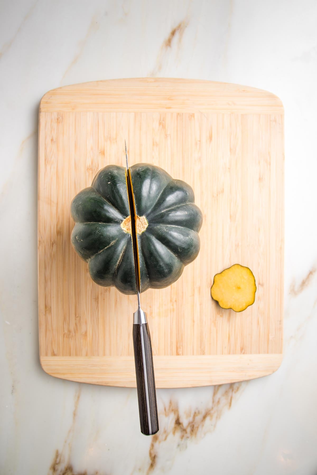 Acorn squash with a sliver of the bottom cut off. THere is a knife inserted through the center stem on top of a cutting board.