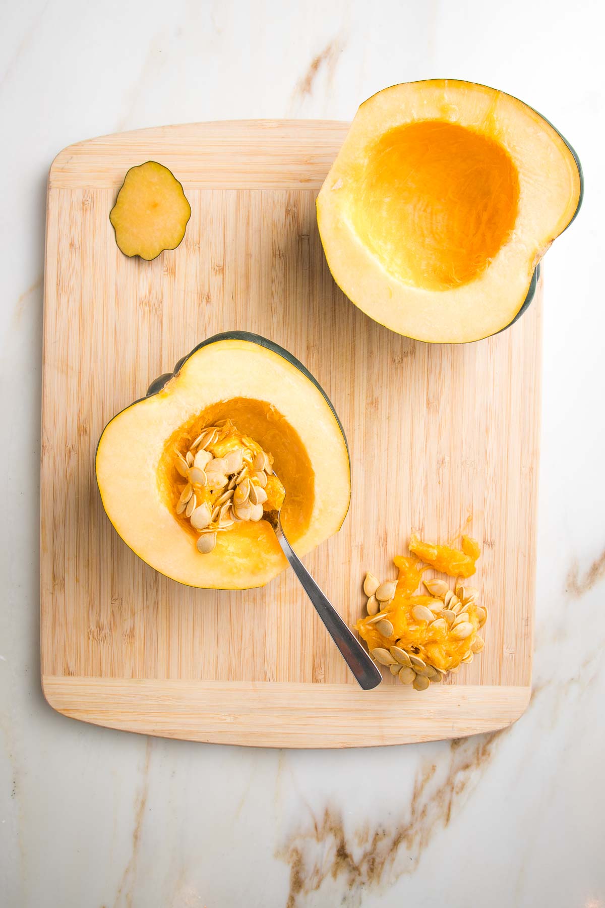 Bamboo cutting board with halved acorn squash. A grapefruit spoon is used to scoop out the seeds and pulp from each half.