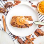 Slice of pumpkin bread with a dollop of almond butter on a white plate. There are dates, cinnamon sticks, pecans and chocolate chips strewn around the sides of the plate.