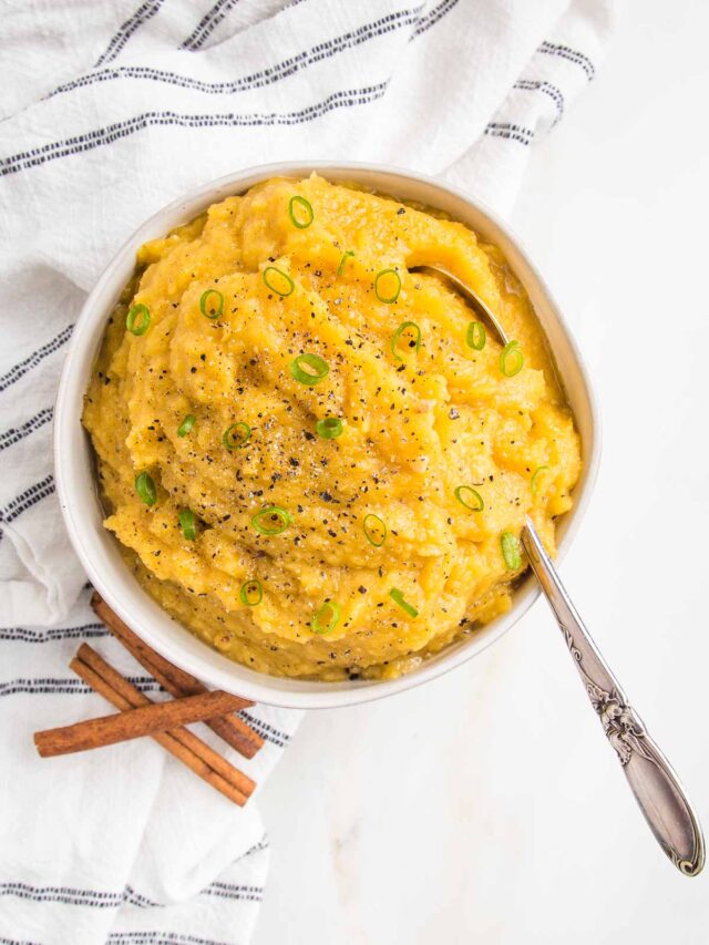 White bowl filled with mashed acorn squash. Garnished with thinly sliced scallions, and styled with a few cinnamon sticks and a navy striped dish towel.