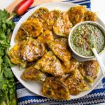 White rimmed round plate filled with appetizer sized baked samosas plus a small white dish filled with green chutney. The plate is on a blue patterned napkin and is surrounded by a bunch of cilantro and a red chili pepper