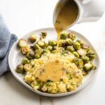 White pitcher pouring gravy over plate of chunky mashed potatoes and brussels sprouts