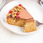 A slice of pumpkin cake with chocolate frosting, candied pepitas and pomegranate seeds. Fork has sectioned off one bite which looks moist and delicious.