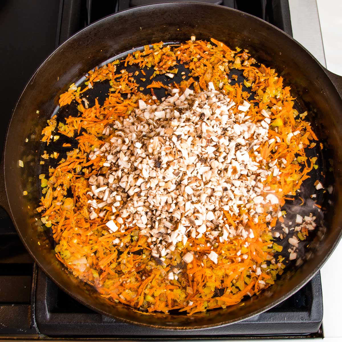 Cast iron skillet with sauteed veggies including shredded carrots. Small diced mushrooms are added to the top of the pan.