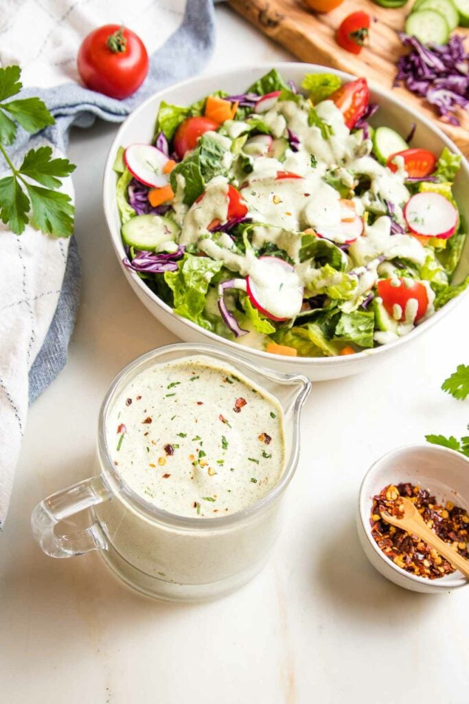 Vegan Ranch dressing in a glass pitcher and also seen drizzled over a simple green salad in the background.