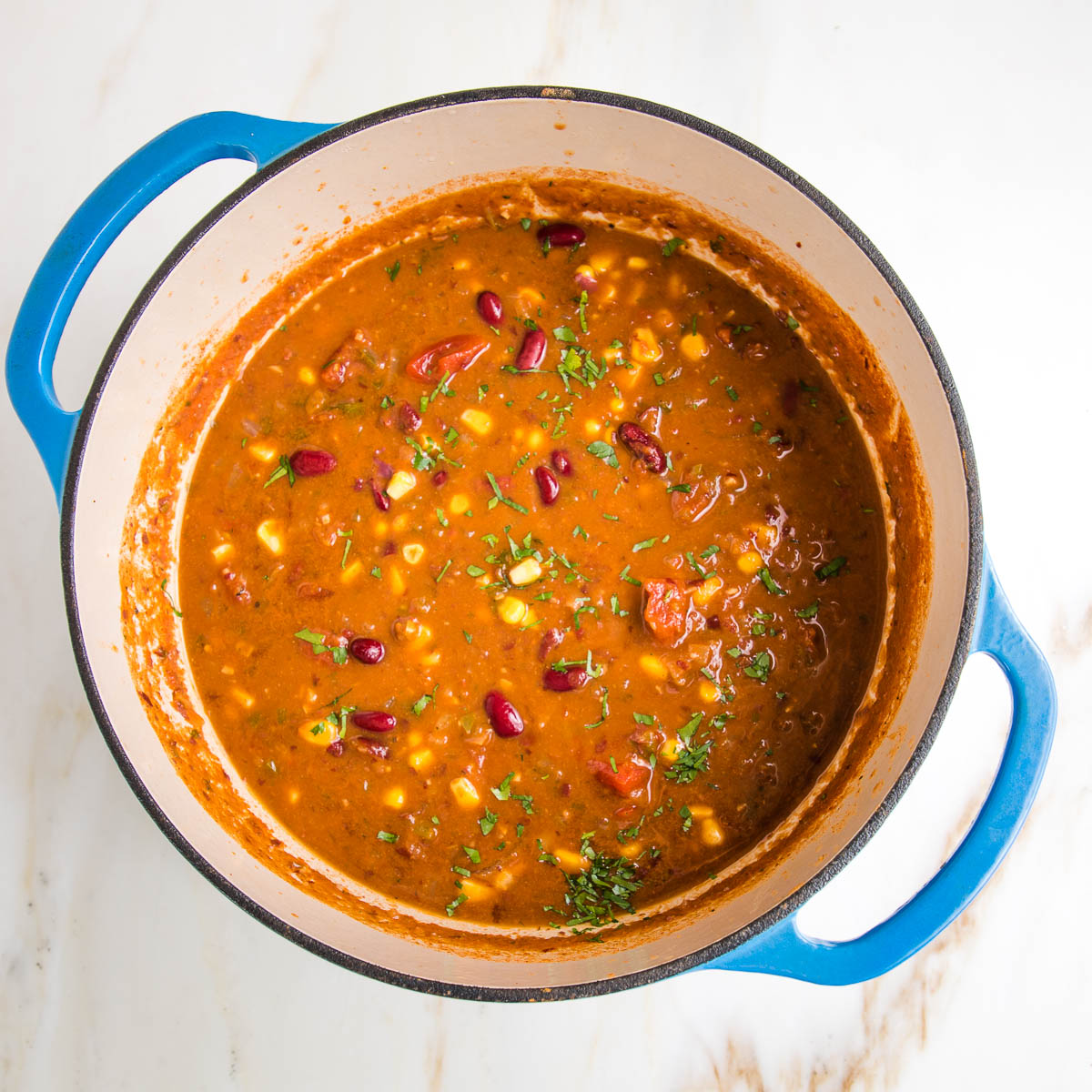 colorful taco soup in a blue dutch oven. Tomato base and you can see red kidney beans, fresh chopped cilantro, corn, pieces of tomatoes and onions.