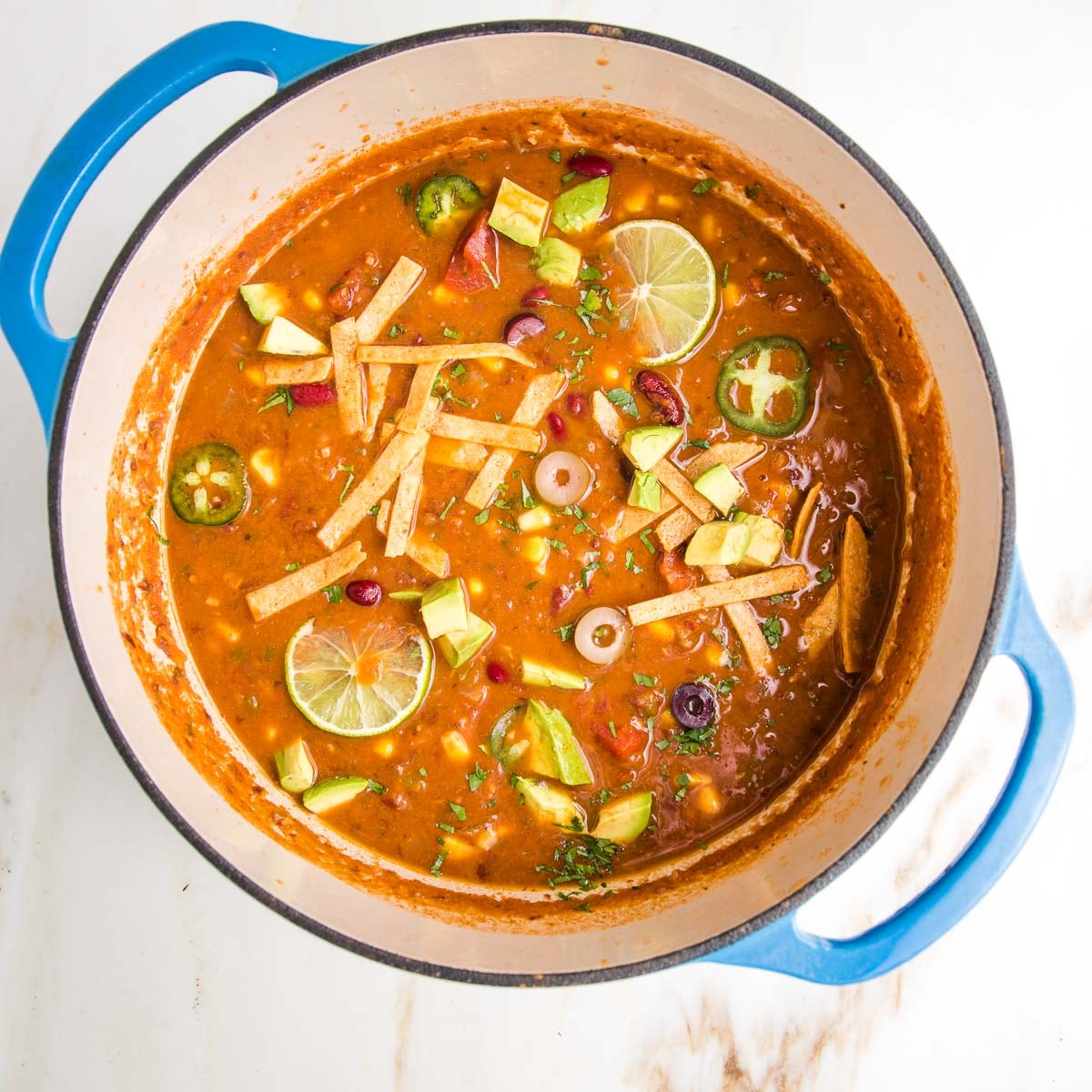 Bright and vibrant vegan tortilla soup. Dutch oven is filled with tomato based soup and decorated with chunks of avocado, lime wedges, sliced jalapenos and olives, plus baked tortilla strips.
