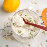 glass jar filled with vegan ricotta, sprinkled with fresh herbs...a spoon is scooping some out. THere are slivered almonds, garlic cloves and a lemon in the background.