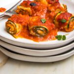 Stack of white plates. On top is a plate full of zucchini roll ups filled with vegan ricotta and topped with marinara.