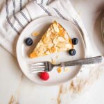 small white plate with slice of lemon cake topped with sliced almonds. Plate has a few blueberries, a raspberry and some almond slices.