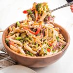 wooden bowl filled with penne pasta and lots of colorful veggies. There is a large spoon scooping up a bunch to plate.