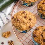 zucchini muffin with chocolate chips, cooling on a wire rack. There is a fresh zucchini on a tea towel in the background along with a couple walnuts and a chocolate chip.
