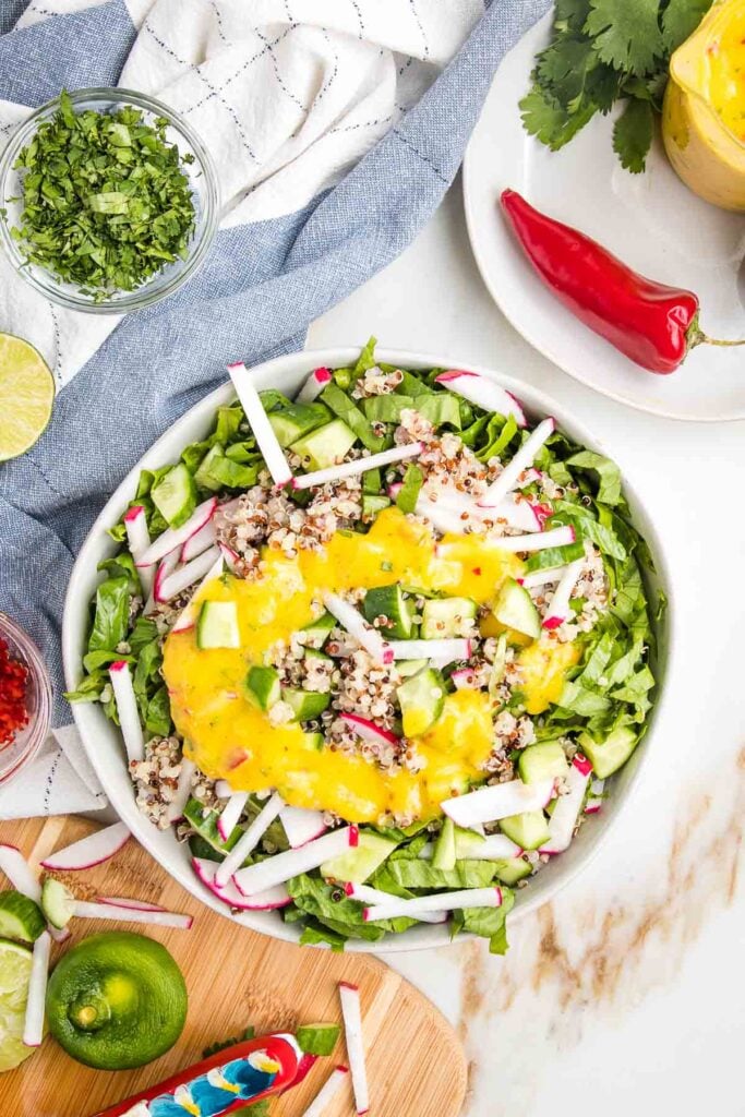 Large white salad bowl filled with julienned romaine, radishes, cucumbers, drizzled with bright yellow mango salad dressing. Dispersed on the counter are cilantro, limes, red hot chili peppers and chunks of veggies.