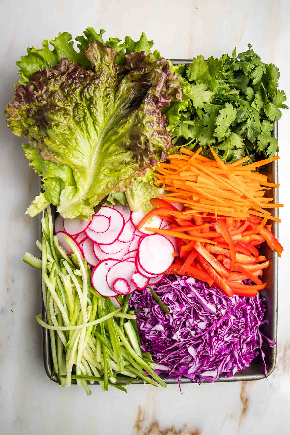 Spring roll ingredients prepped on a tray. Fresh lettuce leaves and cilantro. Thinly sliced radish, shredded cabbage, julienned carrots, red peppers and cucumbers.