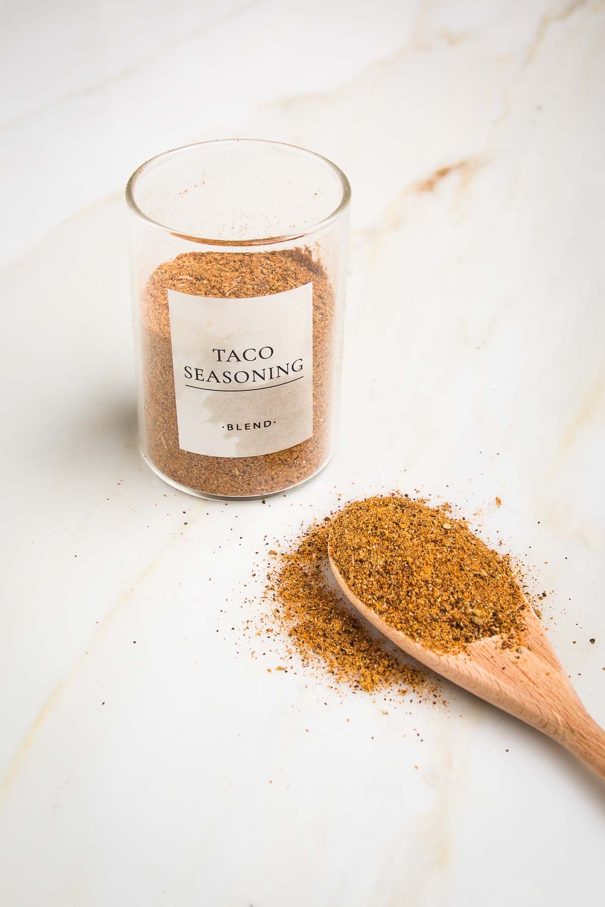 Small glass jar labeled Taco Seasoning Blend ¾ filled with orangish brown powder and a wooden spoon in front of it with same seasoning.