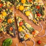 Flatbread Pizza with pesto, hummus, yellow and red tomatoes plus fresh basil and red onions. One slice shown up close and a few others partially shown on a wooden cutting board.