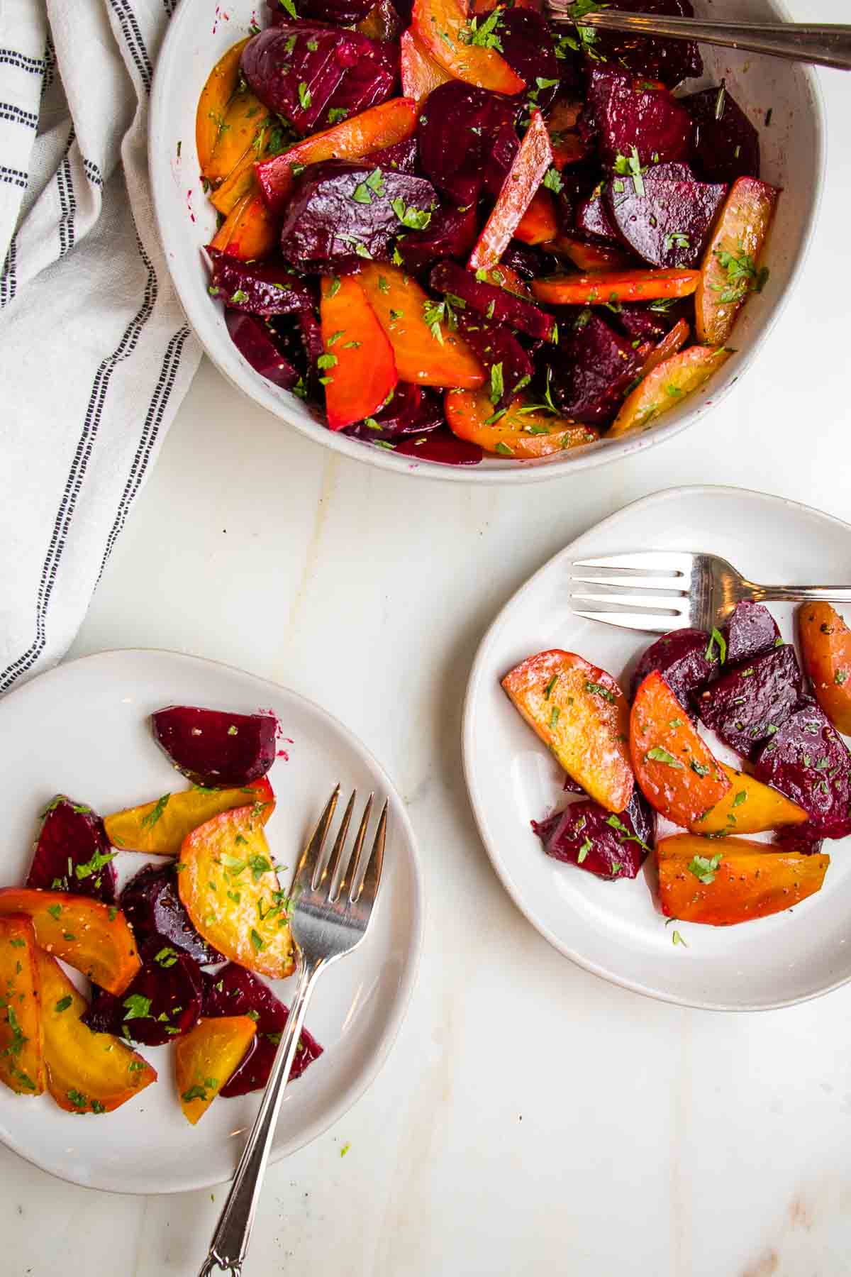 Roasted beet salad sprinkled with chopped parsley in a white bowl. Two small plates with servings of beet salad.