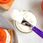 Vegan mayo in a glass jar. Purple spreader plus bright red tomatoes surround it.